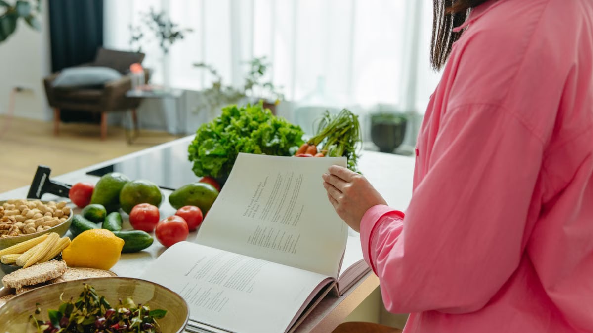 A woman in a kitchen flipping through a cookbook surrounded by fresh vegetables.
