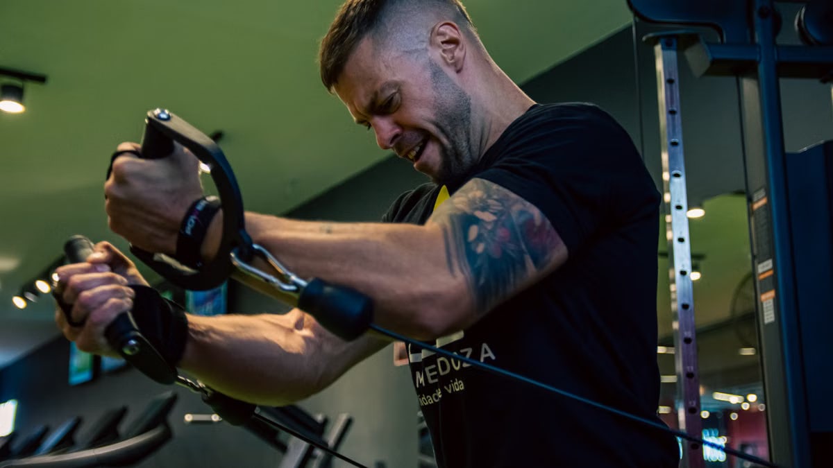 Muscular man with tattoos exercising on a cable machine in a gym, showcasing strength and determination.